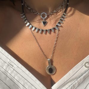 Può includere: Un set di tre collane d'argento. La collana superiore ha un design a catena con una chiusura circolare e un piccolo ciondolo triangolare nero. La collana centrale ha un design a punte. La collana inferiore ha un ciondolo circolare con una pietra scura.