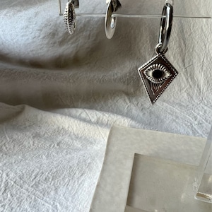 May include: Three silver earrings on a display stand. One earring is a simple hoop, another is a small hoop with a textured surface, and the third is a hoop with a diamond-shaped charm featuring an eye design.