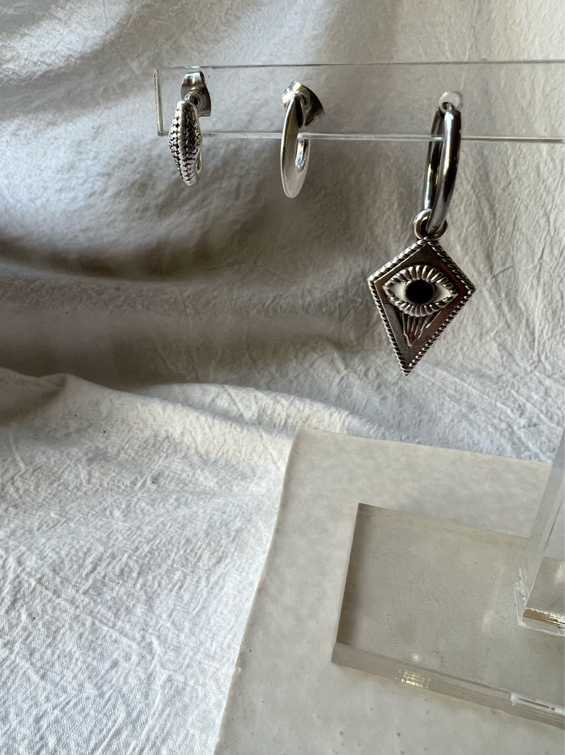 May include: Three silver earrings on a display stand. One earring is a simple hoop, another is a small hoop with a textured surface, and the third is a hoop with a diamond-shaped charm featuring an eye design.