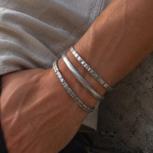Op de afbeelding: Een set van drie zilverkleurige metalen armbanden gedragen om de pols. De armbanden hebben verschillende texturen: één heeft een glad, afgerond oppervlak, terwijl de andere twee ingewikkelde, gesegmenteerde en geëtste patronen vertonen.