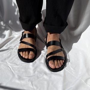 K&ouml;nnte beinhalten: Schwarze Ledersandalen mit mehreren Riemen und Schnallenverschl&uuml;ssen. Die Sandalen haben ein minimalistisches Design mit einer wei&szlig; gen&auml;hten Sohle. Das Bild zeigt die Sandalen aus der Vogelperspektive.