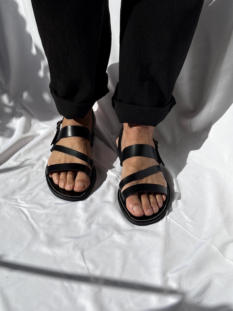 K&ouml;nnte beinhalten: Schwarze Ledersandalen mit mehreren Riemen und Schnallenverschl&uuml;ssen. Die Sandalen haben ein minimalistisches Design mit einer wei&szlig; gen&auml;hten Sohle. Das Bild zeigt die Sandalen aus der Vogelperspektive.