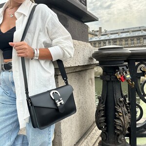 Puede incluir: Una mujer con una camisa blanca, pantalones vaqueros azules y un bolso bandolera de cuero negro con una hebilla plateada. El bolso tiene una correa larga y se lleva sobre el hombro.
