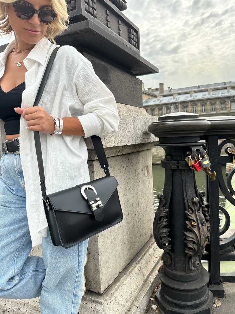 Puede incluir: Una mujer con una camisa blanca, pantalones vaqueros azules y un bolso bandolera de cuero negro con una hebilla plateada. El bolso tiene una correa larga y se lleva sobre el hombro.