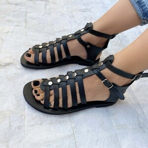 Puede incluir: Un par de sandalias gladiadoras de cuero negro con hebillas y tachuelas plateadas. Las sandalias tienen m&uacute;ltiples correas que se cruzan en el pie y el tobillo.