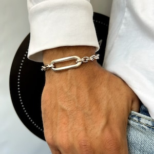 K&ouml;nnte beinhalten: Ein silbernes Kettenarmband mit einem gro&szlig;en ovalen Glied. Das Armband befindet sich am Handgelenk einer Person.