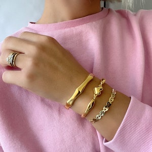 May include: A set of three gold bracelets on a person's wrist. The bracelets are a thick, textured bangle, a thin, curved bangle, and a chain bracelet with a heart charm.