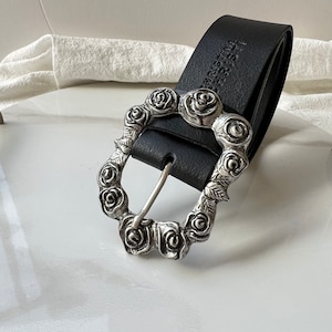 Peut inclure: Ceinture en cuir noir avec une boucle argentée ornée de roses et de feuilles. La ceinture est un accessoire classique. La boucle est un motif décoratif de roses et de feuilles.