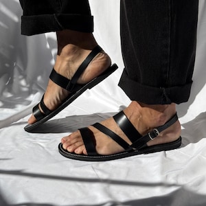 K&ouml;nnte beinhalten: Schwarze Ledersandalen mit mehreren Riemen und einem Schnallenverschluss. Die Sandalen haben eine flache Sohle und werden mit einer schwarzen Hose getragen. Das Bild ist in nat&uuml;rlichem Licht aufgenommen, wodurch die Lederstruktur und das Design der Sandalen hervorgehoben werden.