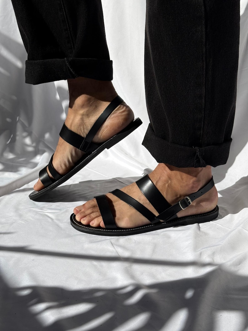 K&ouml;nnte beinhalten: Schwarze Ledersandalen mit mehreren Riemen und einem Schnallenverschluss. Die Sandalen haben eine flache Sohle und werden mit einer schwarzen Hose getragen. Das Bild ist in nat&uuml;rlichem Licht aufgenommen, wodurch die Lederstruktur und das Design der Sandalen hervorgehoben werden.