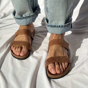 May include: Brown leather sandals with two adjustable straps. The sandals have a cork footbed and a black sole. The person is wearing light blue denim jeans with the cuffs rolled up.