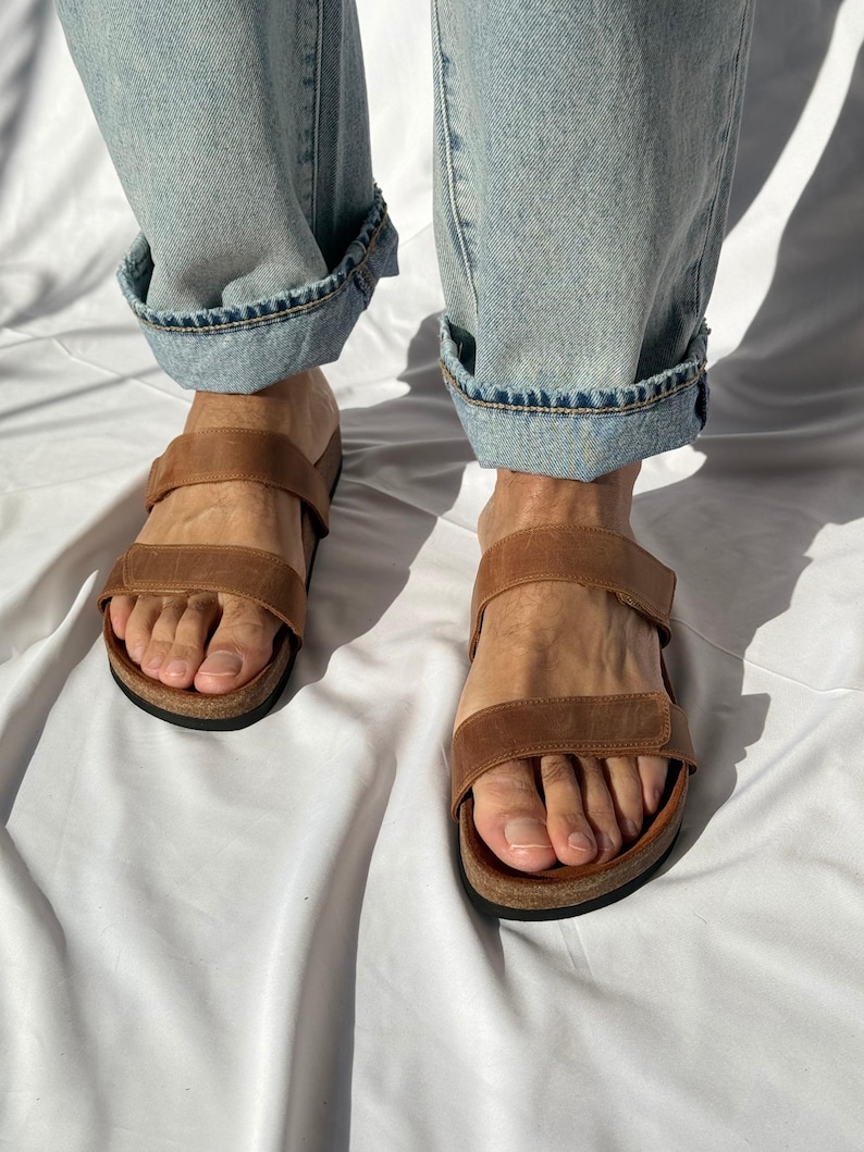 May include: Brown leather sandals with two adjustable straps. The sandals have a cork footbed and a black sole. The person is wearing light blue denim jeans with the cuffs rolled up.