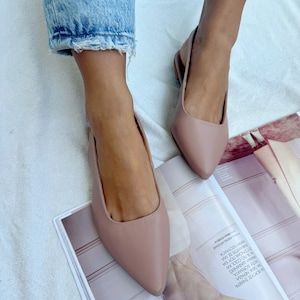 Könnte beinhalten: Ein Paar hellrosa spitze Slipper mit Riemchen am Knöchel. Die Schuhe sind aus weichem, glattem Leder gefertigt und bieten einen bequemen Sitz.