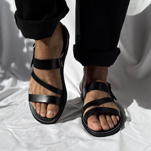 Op de afbeelding: Zwarte leren sandalen met meerdere bandjes en een gespsluiting. De sandalen hebben een wit gestikte zool. De persoon draagt een zwarte broek met opgerolde manchetten.
