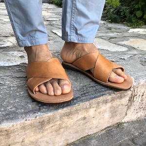 May include: A pair of brown leather sandals with a criss-cross strap design. The sandals are worn on a person's feet, with the toes visible.