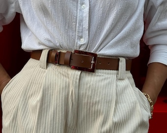May include: A white button-down shirt with a brown leather belt featuring a red and clear plastic buckle. The belt is worn over beige corduroy trousers.