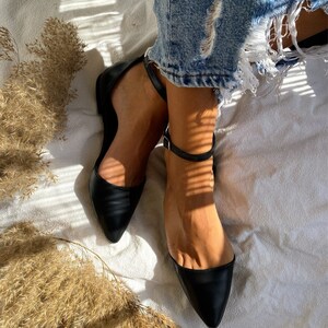 May include: A pair of black leather ankle strap flats with pointed toes. The shoes are worn by a person sitting on a white surface.