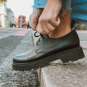May include: Black leather lace-up shoes with a thick platform sole. The shoes are being tied by a person wearing blue jeans and a red and white striped shirt.