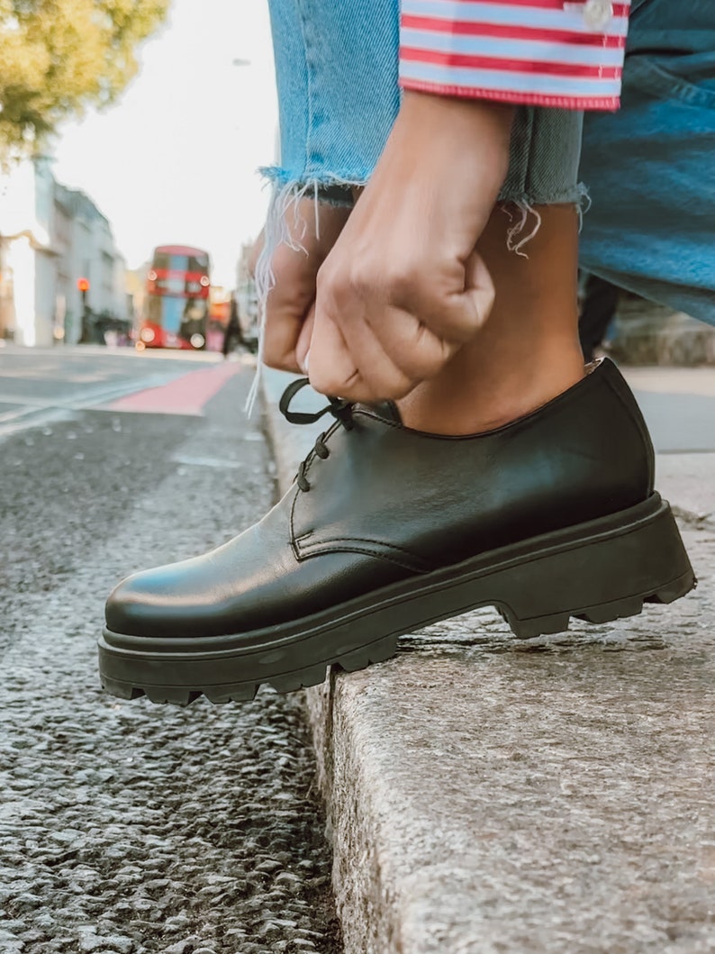 May include: Black leather lace-up shoes with a thick platform sole. The shoes are being tied by a person wearing blue jeans and a red and white striped shirt.