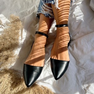 May include: Black pointed-toe flats with ankle straps. The shoes have a sleek, minimalist design, with a smooth, leather-like texture. The ankle straps are thin and adjustable, adding a touch of elegance. The flats are set against a soft, white background.