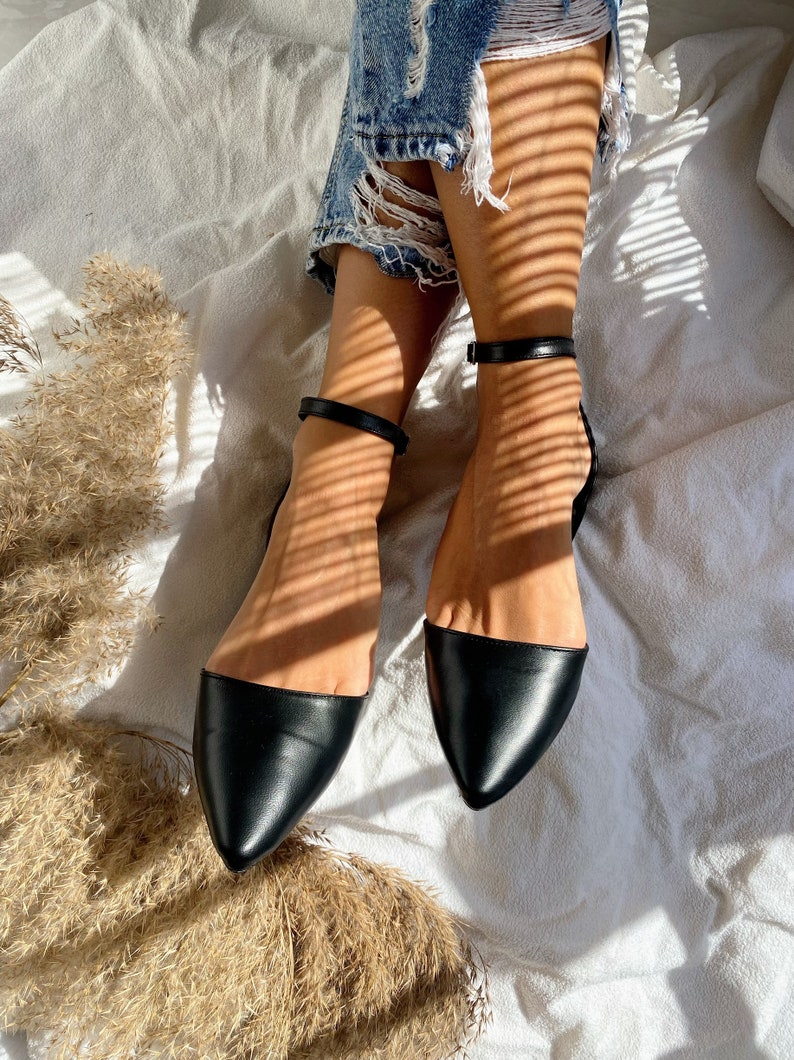 May include: Black pointed-toe flats with ankle straps. The shoes have a sleek, minimalist design, with a smooth, leather-like texture. The ankle straps are thin and adjustable, adding a touch of elegance. The flats are set against a soft, white background.