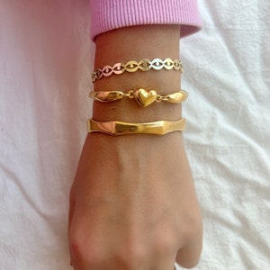 May include: A set of three gold bracelets, one with a heart charm, one with a chain link design, and one with a thick, textured band.