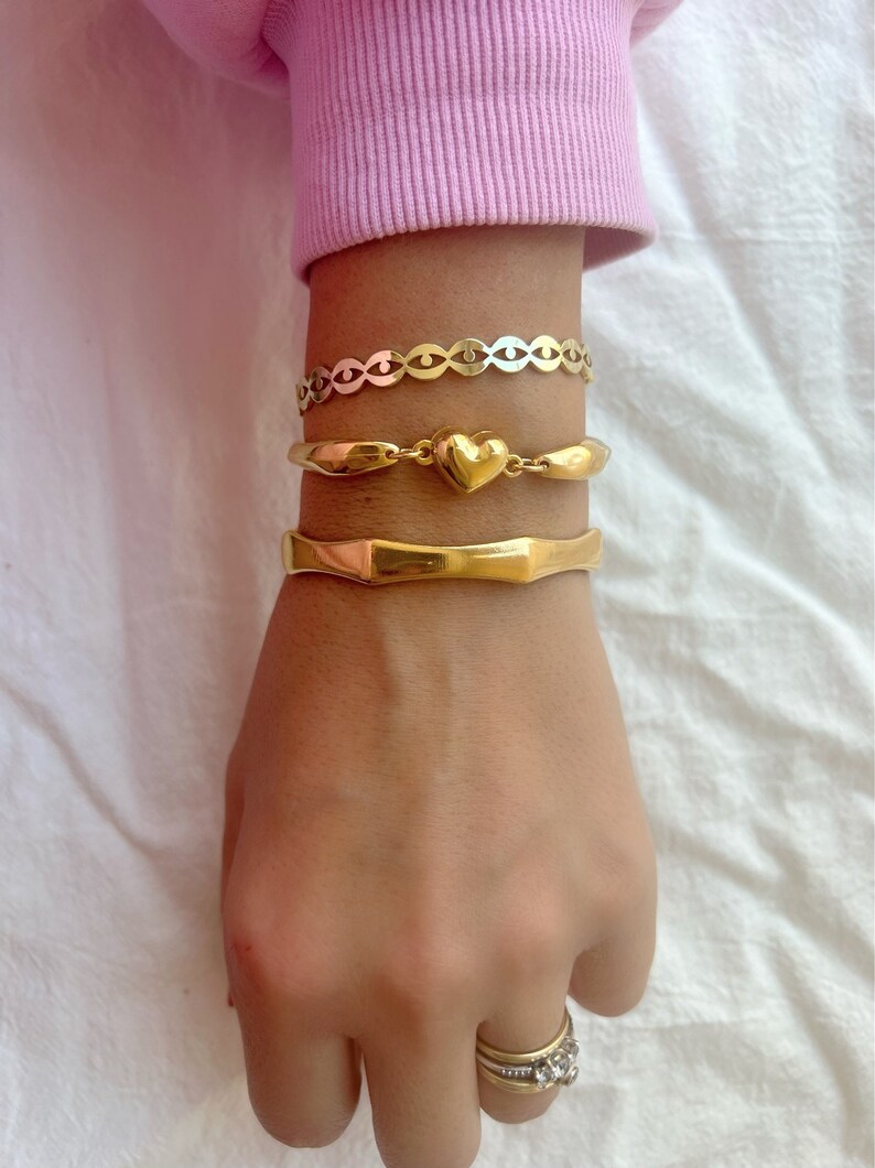 May include: A set of three gold bracelets, one with a heart charm, one with a chain link design, and one with a thick, textured band.