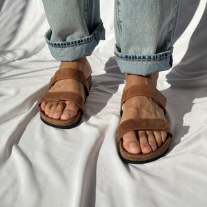 May include: Brown leather sandals with two adjustable straps and a cork footbed. The sandals are worn with light blue denim jeans with rolled-up cuffs. The sandals are designed for comfort and style.