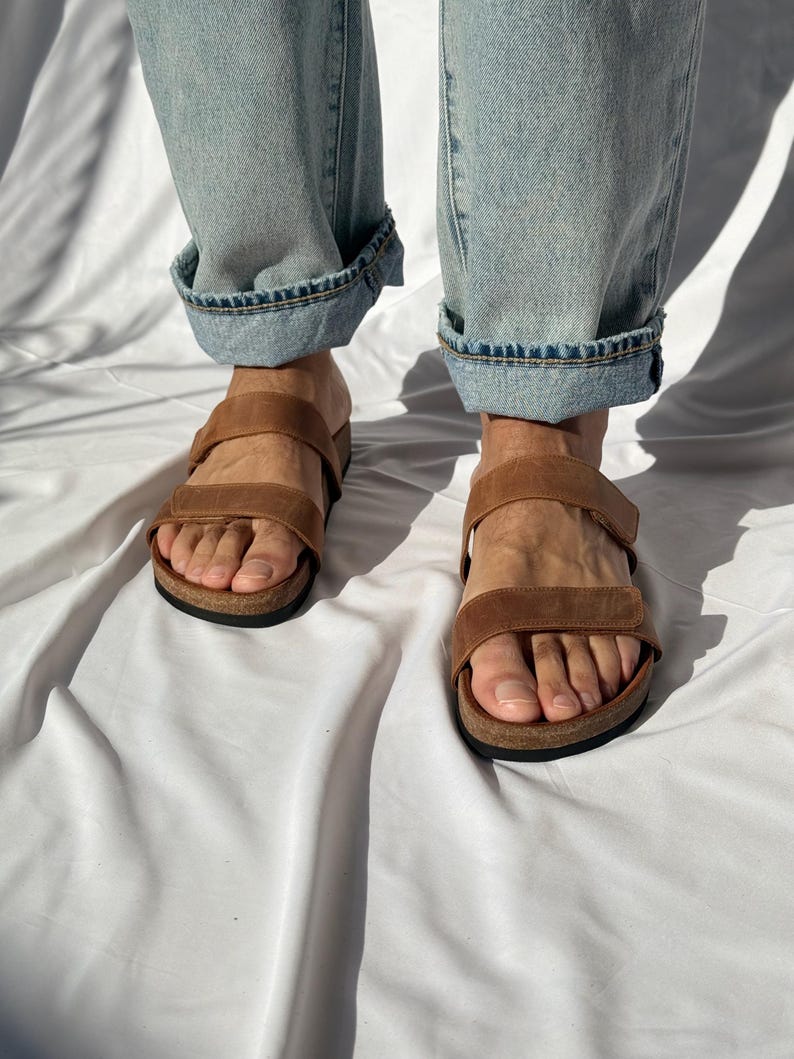 May include: Brown leather sandals with two adjustable straps and a cork footbed. The sandals are worn with light blue denim jeans with rolled-up cuffs. The sandals are designed for comfort and style.