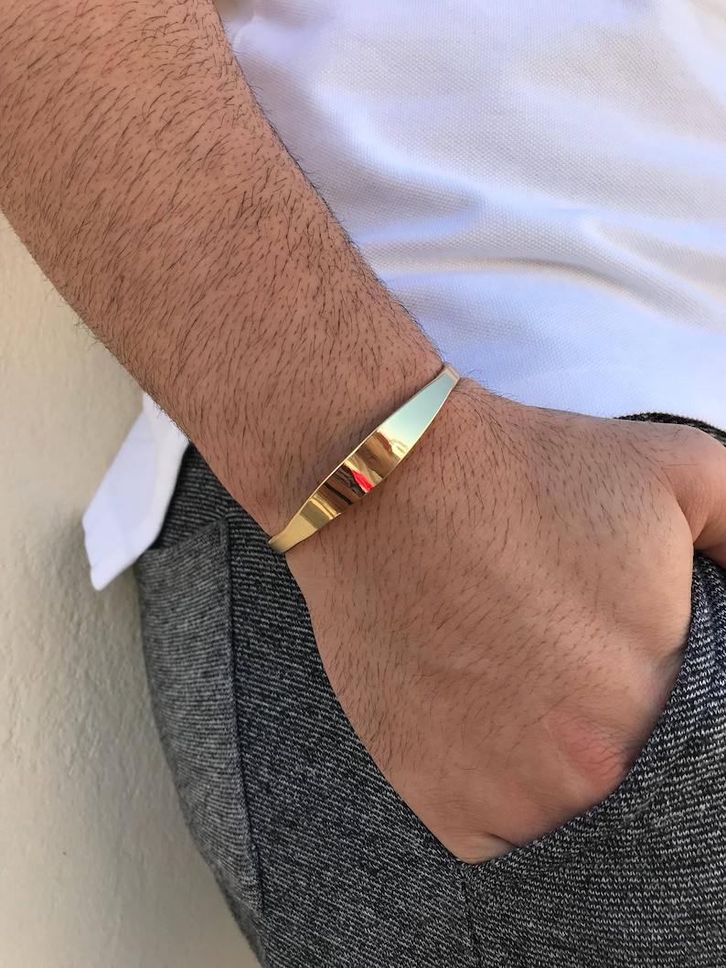 May include: A gold-toned metal bracelet with a flat, slightly curved design. The bracelet is worn on a person's wrist.
