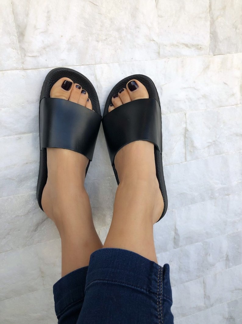 May include: A pair of black leather slides with a simple design. The slides have a black rubber sole and a black leather strap.