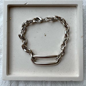 K&ouml;nnte beinhalten: Ein silbernes Kettenarmband mit einem gro&szlig;en, ovalen Glied. Das Armband besteht aus einer dicken, massiven Kette und hat ein schlichtes, modernes Design.