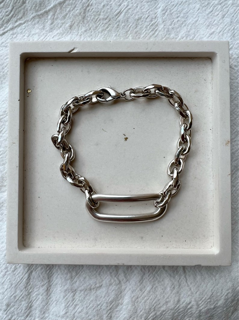 K&ouml;nnte beinhalten: Ein silbernes Kettenarmband mit einem gro&szlig;en, ovalen Glied. Das Armband besteht aus einer dicken, massiven Kette und hat ein schlichtes, modernes Design.
