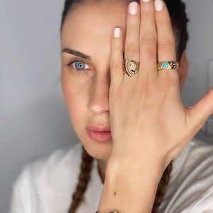 May include: A woman's hand with three gold rings on her finger. The rings feature an eye design, a turquoise stone, and a geometric pattern. She is wearing a silver and gold watch on her wrist.