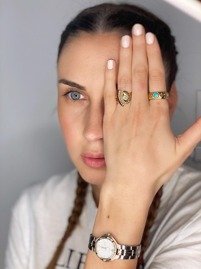 May include: A woman's hand with three gold rings on her finger. The rings feature an eye design, a turquoise stone, and a geometric pattern. She is wearing a silver and gold watch on her wrist.