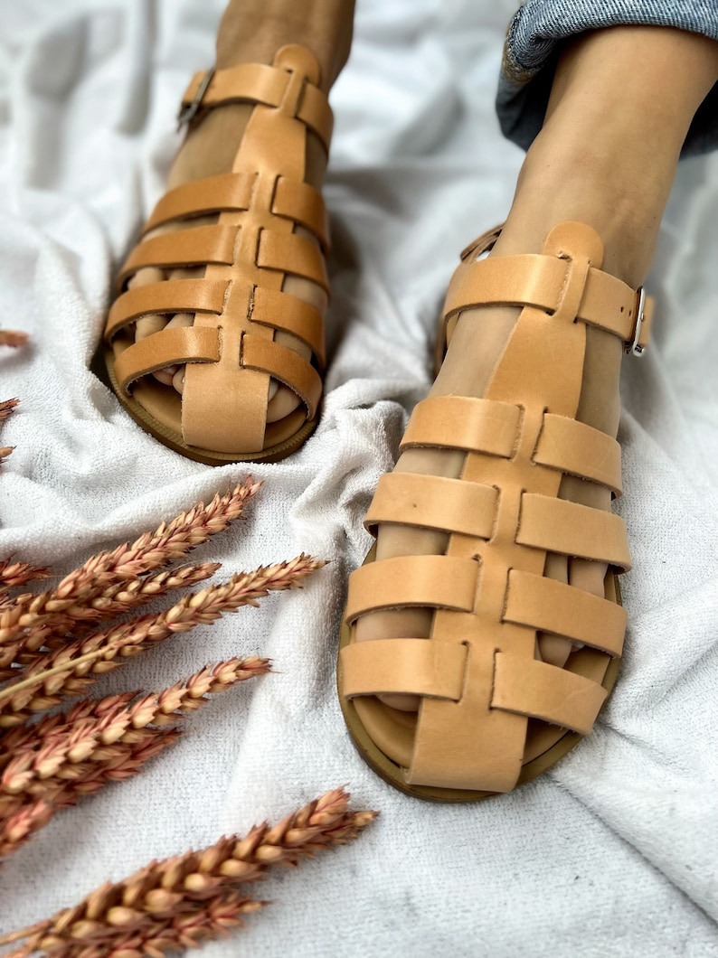 Op de afbeelding: Een paar bruine leren sandalen met een geweven ontwerp. De sandalen hebben een gespsluiting en een platte zool.