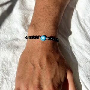 May include: A black beaded bracelet with a silver charm featuring a blue evil eye.