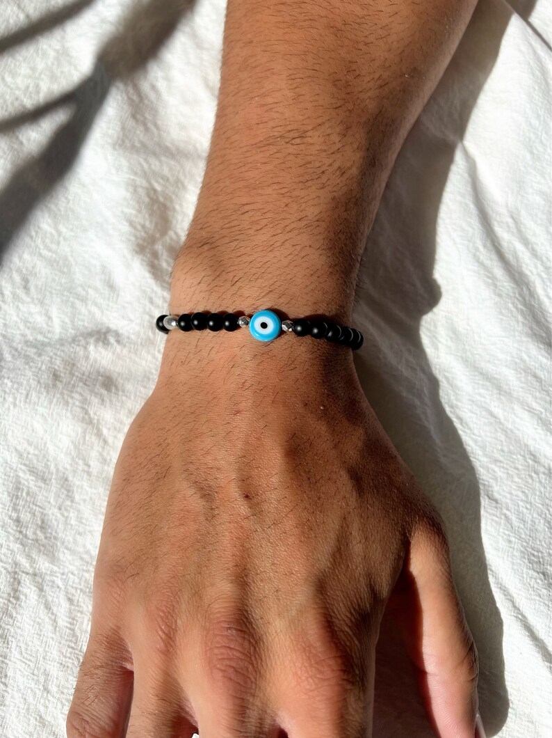 May include: A black beaded bracelet with a silver charm featuring a blue evil eye.