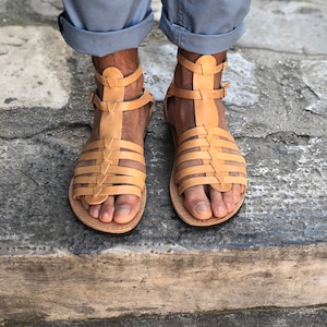 Puede incluir: Un par de sandalias gladiador de cuero color arena con m&uacute;ltiples correas y hebillas. Las sandalias se llevan en los pies de una persona.