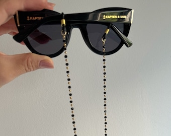 Könnte beinhalten: Schwarze Sonnenbrille mit einer goldenen Kette und dem Text "KAPTEN & SON" auf der rechten Linse.
