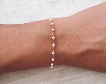 Puede incluir: Delicada pulsera dorada con pequeñas perlas blancas ensartadas en una fina cadena dorada. La pulsera está diseñada para llevarse alrededor de la muñeca, mostrando una pieza de joyería minimalista y elegante.