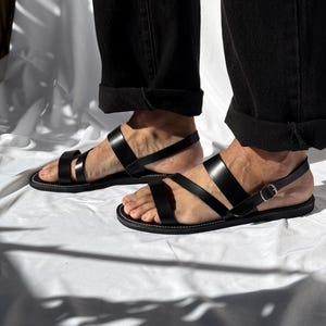 K&ouml;nnte beinhalten: Schwarze Ledersandalen mit mehreren Riemen und einem Schnallenverschluss. Die Sandalen haben ein minimalistisches Design mit wei&szlig;en N&auml;hten um die Sohle. Das Bild zeigt die Sandalen auf einer wei&szlig;en Oberfl&auml;che.