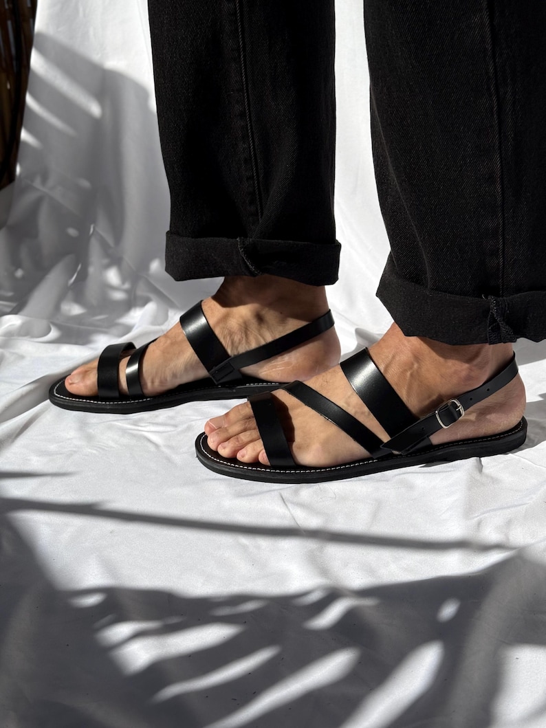 K&ouml;nnte beinhalten: Schwarze Ledersandalen mit mehreren Riemen und einem Schnallenverschluss. Die Sandalen haben ein minimalistisches Design mit wei&szlig;en N&auml;hten um die Sohle. Das Bild zeigt die Sandalen auf einer wei&szlig;en Oberfl&auml;che.