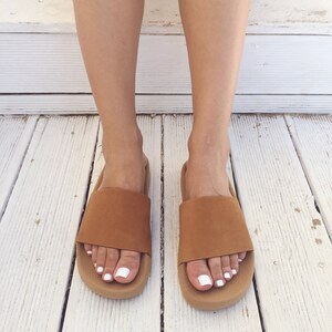 May include: A pair of brown leather slide sandals with a white sole. The sandals are worn on a white wooden surface.