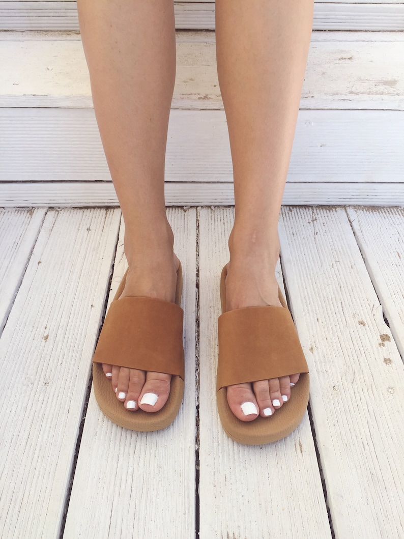 May include: A pair of brown leather slide sandals with a white sole. The sandals are worn on a white wooden surface.