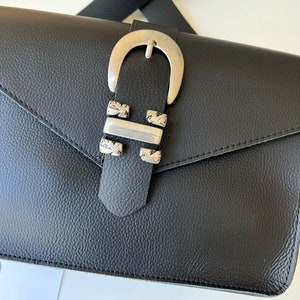 Puede incluir: Bolso bandolera de cuero negro con hebilla plateada y cierre de solapa. El bolso tiene un acabado de cuero texturizado.