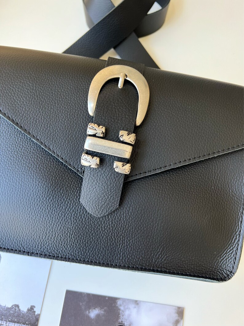 Puede incluir: Bolso bandolera de cuero negro con hebilla plateada y cierre de solapa. El bolso tiene un acabado de cuero texturizado.