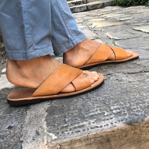 May include: Tan leather sandals with a criss-cross strap design. The sandals have a flat sole and are worn on a person's feet.