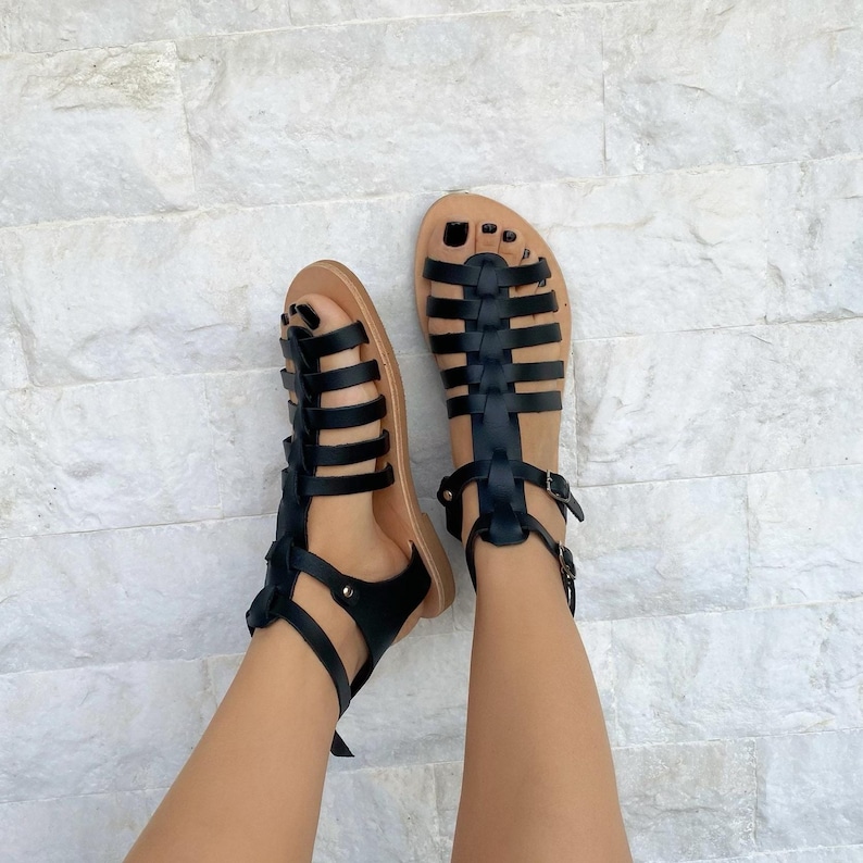 May include: A pair of black gladiator sandals featuring multiple horizontal straps across the foot, a vertical center strap, and an adjustable ankle strap with a small metal buckle. The sandals have a tan-colored footbed and a flat sole design.