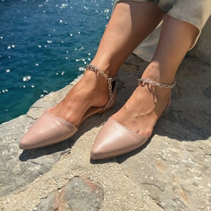May include: A pair of blush-colored pointed-toe flats with silver chain anklets. The flats are made of a smooth material and have a low-cut vamp. The anklets feature a chunky chain design.
